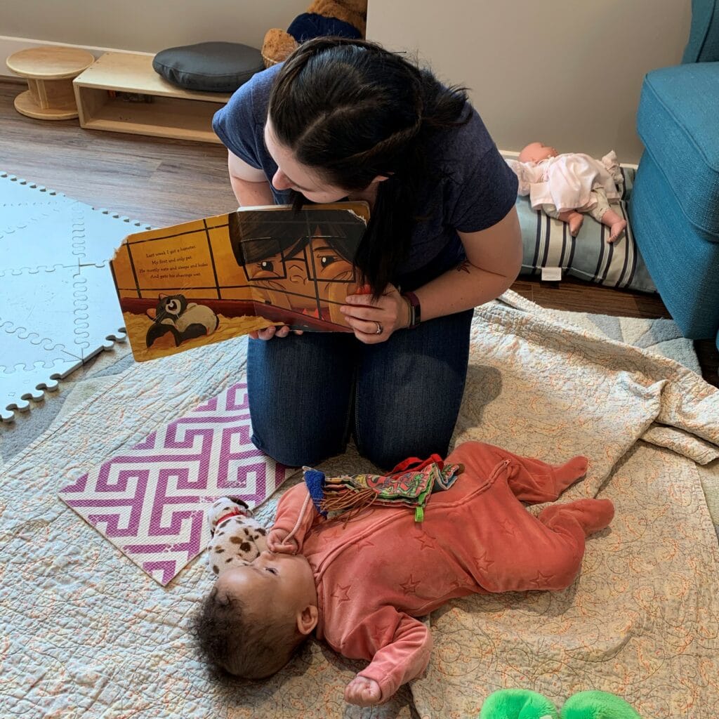 Responsive infant teacher reading with young baby on a blanket.