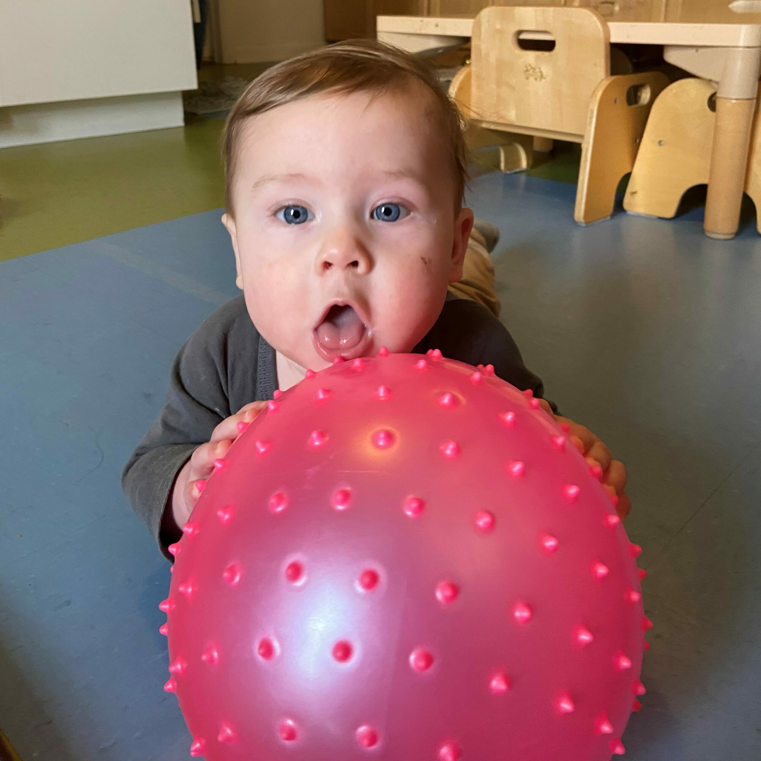 Infant grabs a pink/red ball and sticks tongue out, making focused eye contact
